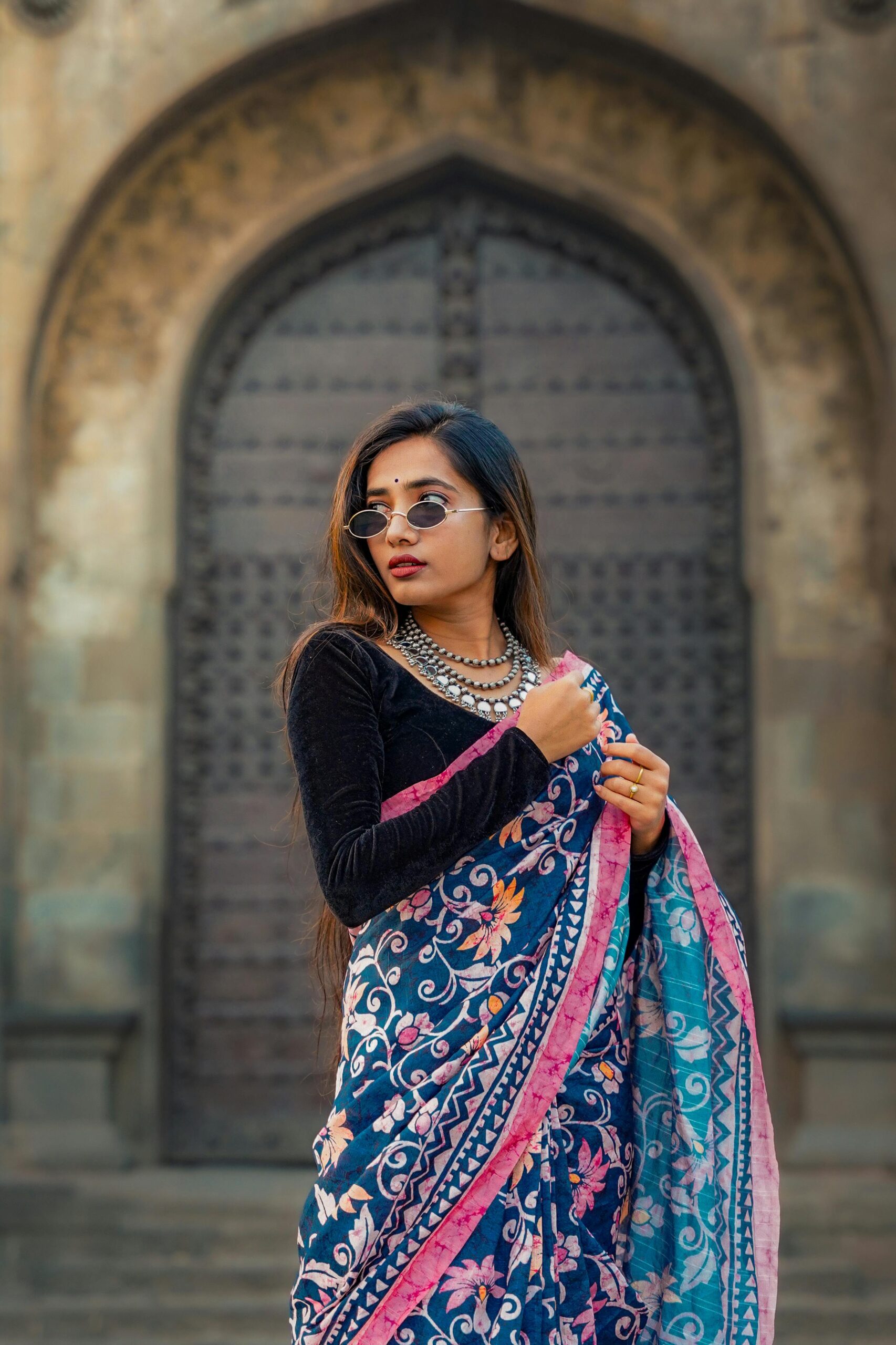 Stylish Indian woman poses in colorful saree and jewelry against an architectural backdrop in Pune.